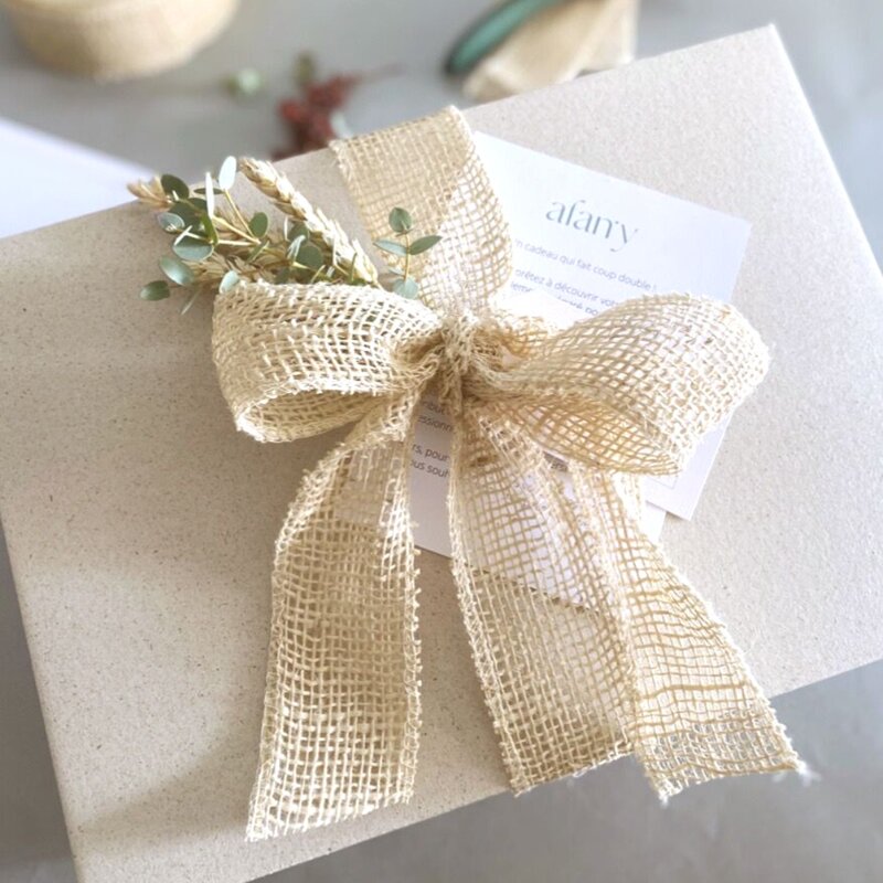 L'image représente un coffret en papier d'herbe fermé par ruban en toile de jute de couleur claire. Le coffret est agrémenté de branches d'eucalyptus et de blé. Il est accompagné d'une carte