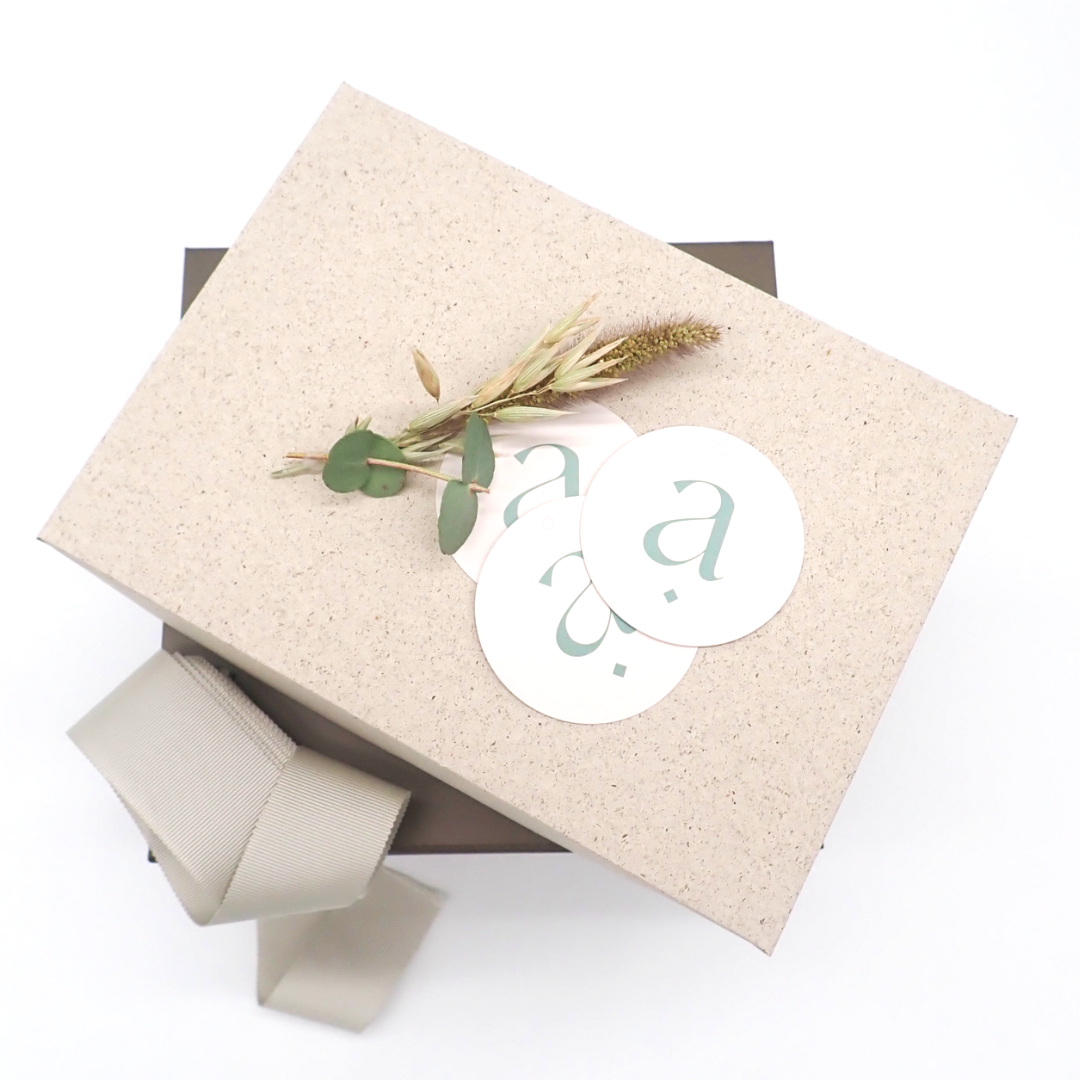 L'image représente un coffret en papier d'herbe posé sur un coffret aimanté. Sur le coffret du haut sont disposés 3 autocollants avec le logo afan'y ainsi qu'un petit bouquet de fleurs séchées. Un ruban de couleur claire est posé sur le côté des coffrets.